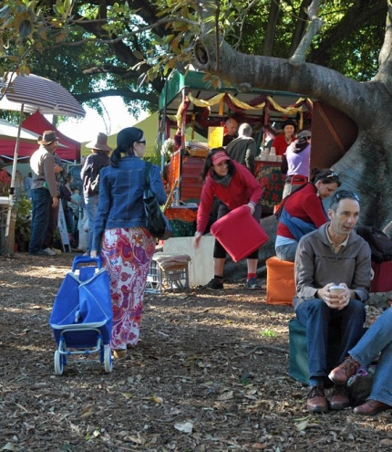 People sitting under a tree in the Davies Park Market with stalls in the background.