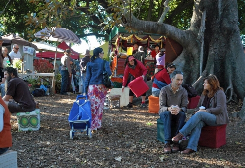 People sitting under a tree in the Davies Park Market with stalls in the background.